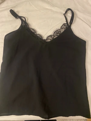 Svart linnetopp med spetsdetalj! - Säljer en svart linnetopp med tunna justerbara axelband och snygg spetskant vid v-ringningen. Toppen har en enkel och stilren design som är lätt att matcha med olika outfits. Materialet känns mjukt och har en lätt glans. Använd 1 gång!