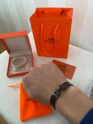 Svart Hermès skinnarmband med H-spänne - Säljer ett stilrent svart armband från Hermès med klassiskt H-spänne i metall. Armbandet är tillverkat i skinn och kommer med original ask, påse och certifikat. Perfekt accessoar för dig som gillar lyxiga detaljer och ikonisk design.