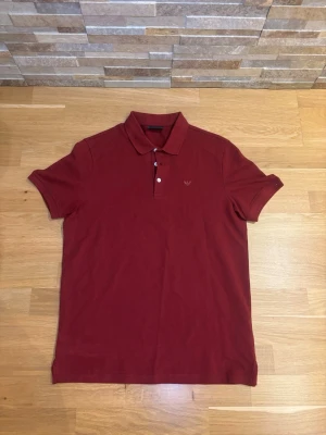 Mörkröd piké från Emporio Armani - Säljer en stilren mörkröd pikétröja från Emporio Armani med klassisk krage och tre vita knappar framtill. Pikén har korta ärmar och är tillverkad i mjuk bomull. På bröstet finns det ikoniska Armani-logotypen broderad ton-i-ton.