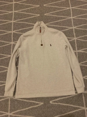 Beige half zip tröja från Polo Ralph Lauren - Säljer en beige långärmad tröja från Polo Ralph Lauren i storlek M. Tröjan har en half zip-dragkedja med läderdetalj och klassisk broderad logga på bröstet. Materialet är mjukt och känns som bomull, perfekt för kyliga dagar.