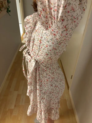 Blommig kortklänning med volang - Supersöt kortklänning med blommigt mönster i rosa, gul och grönt på vit botten. Klänningen har puffiga korta ärmar, volangdetaljer längs nederkanten och omlottkänsla med knyt i midjan. Dragkedja bak för enkel påklädning. Perfekt för sommarens alla tillfällen!