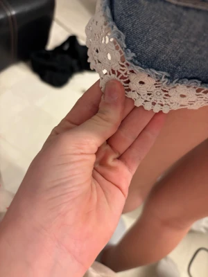 Blå jeansshorts med spetskant - Säljer ett par blå denimshorts med snygga slitningar och vit spetskant längst ner. Shortsen har klassisk femficksdesign, bälteshällor och knappstängning. Perfekta för varma dagar och ger en söt touch med spetsdetaljen.