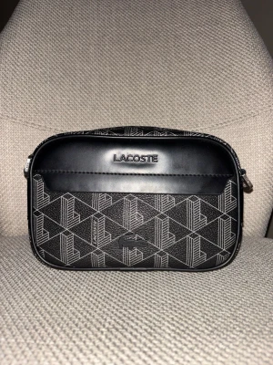 Lacoste Väska - Snygg svart axelremsväska från Lacoste med geometriskt vitt mönster och silverfärgade detaljer. Väskan har justerbar axelrem, dragkedja och präglad Lacoste-logga framtill. Tillverkad i syntetmaterial med stilren design och praktiskt ytterfack. Köpt för 1750kr 