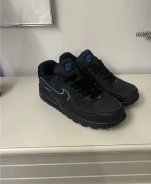 Nike Air Max 90 svart/blå sneakers - Säljer ett par svarta Nike Air Max 90 sneakers med blå detaljer och logga. Skorna har en klassisk siluett, snörning och en synlig Air Max-dämpning i sulan. Ovandelen är i en mix av textil och syntet med snygga kontrasterande blå swoosh och detaljer.