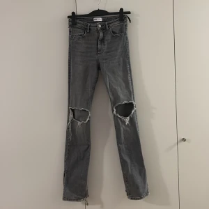 Grå slitna jeans med slits från Zara - Säljer ett par grå jeans från Zara med snygga slitningar över knäna och slits vid bensluten. Modellen är rak och har klassisk femficksdesign. Jeansen har hög midja och är tillverkade i jeansmaterial med en cool, tvättad look.