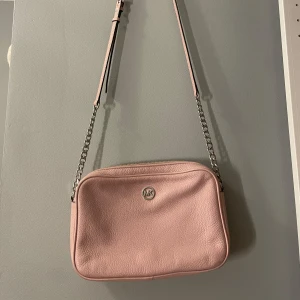 Rosa axelväska från Michael Kors - Säljer en snygg ljusrosa axelväska från Michael Kors i skinn med silverfärgad kedja och justerbar rem. Väskan har ett stilrent MK-emblem på framsidan och dragkedja upptill. Insidan är fodrad med logotypmönster och har praktiska fack för småsaker. Den har lite fläckar på insidan men ska kunnas ta bort flitigt. Ingen syn på användning på utsidan 