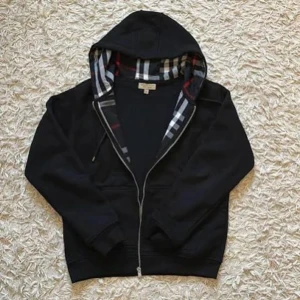 Svart Burberry hoodie med dragkedja - Säljer en svart hoodie från Burberry med klassiskt rutigt foder i huvan. Hoodien har dragkedja framtill, ribbade muddar och fickor på sidorna. Perfekt för dig som gillar stilren design med en touch av lyx. Materialet är mjukt och bekvämt.
