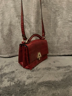 Zara Röd axelväska med krokodilmönster - Snygg röd axelväska från Zara med krokodilmönstrad yta och guldfärgat lås framtill. Väskan har både handtag och axelrem, vilket gör den enkel att bära på olika sätt. Materialet är skinnimitation och designen är stilren med flera fack för smart förvaring.