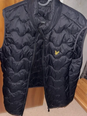 Svart dunväst från Lyle & Scott - Svart dunväst från Lyle & Scott med gul logga på bröstet. Västen har quiltad design med vågiga sömmar och dragkedja framtill. Perfekt att slänga över en hoodie för extra värme. Materialet är polyester och dun, vilket gör den lätt och skön. Finns ett litet litet hål, se bild annars mycket fin!