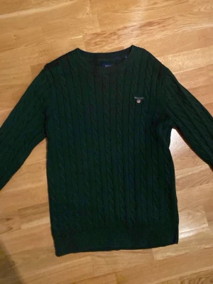 Mörkgrön kabelstickad tröja GANT - Snygg mörkgrön kabelstickad tröja från GANT med rund hals och diskret logga broderad på bröstet. Tröjan har långärmade ribbade muddar och är gjord i mjuk bomull. Perfekt för dig som gillar klassisk stil med en modern touch.