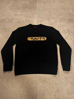 Svart sweatshirt från Kufson med gul detalj - Svart sweatshirt från Kufson med lång ärm och rund hals. På bröstet finns en gul, avlång detalj med svart broderad text. Tröjan har ribbade muddar vid ärmslut och nederkant. Perfekt för dig som gillar stilrena och grafiska prints.