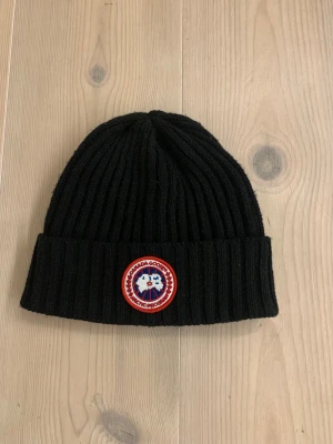 Svart ribbad mössa från Canada Goose - Svart ribbstickad mössa från Canada Goose med klassisk rund logotyp-patch framtill i rött, vitt och blått. Mössan har uppvikt kant och är tillverkad i mjukt stickat material som håller dig varm under kalla dagar. 