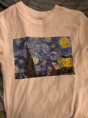 Vit t-shirt med Svampbob och Starry Night - Vit t-shirt med tryck av Svampbob Fyrkant som målar i en tolkning av Van Goghs Starry Night. T-shirten har rund hals och korta ärmar. Materialet känns som bomull och motivet är färgglatt och unikt.