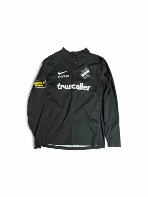 Svart AIK Nike träningsjacka - Svart långärmad träningsjacka från Nike med AIK-logga på bröstet och flera sponsorer tryckta i vitt och gult. Jackan har halv dragkedja framtill och är tillverkad i 100% polyester. Perfekt för dig som gillar fotboll och vill ha en snygg sportig stil.