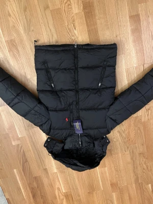 Svart pufferjacka från Polo Ralph Lauren - 🚨🚨Fejk🚨🚨Svart pufferjacka från Polo Ralph Lauren med huva och två snedställda fickor med dragkedja.Jackan har quiltad design och klassisk Polo-logga i rött på bröstet. Perfekt för kalla dagar och har en clean, modern look.