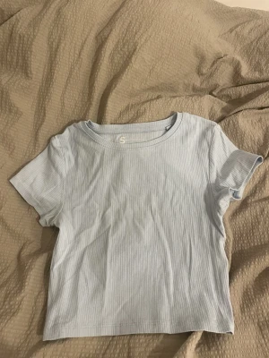 Ljusblå ribbad t-shirt i bomull - Säljer en ljusblå ribbad t-shirt i mjuk bomull. T-shirten har korta ärmar och en klassisk rund halsringning. Perfekt för dig som gillar enkel och fräsch stil med lite struktur i tyget.
