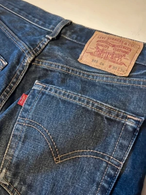Levi's 501 mörkblå jeans med rå kant - Säljer ett par klassiska Levi's 501 jeans i mörkblå tvätt med raka ben och fransig, rå kant nedtill. Jeansen har fem fickor och den ikoniska läderpatchen bak i midjan. Perfekt för dig som gillar tidlös denim med lite edge. Liten i storlek.