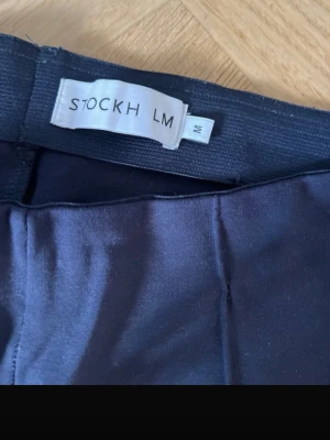 Mörkblå kostymbyxor från STOCKH LM - Snygga mörkblå kostymbyxor från STOCKH LM i storlek M. Byxorna har raka ben, pressveck och diskreta fickor bak. Materialet är mjukt och har en lätt glans. Dragkedja vid benslut för extra stilpoäng.