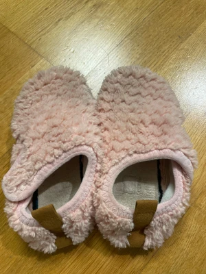 Rosa fluffiga tofflor med gummisula - Supermysiga rosa tofflor med fluffigt yttre och svart gummisula med mönster för extra grepp. Insidan är mjuk och skön, perfekt för att chilla hemma. Tofflorna har en rund tå och en liten brun detalj baktill.- storlekn är lite oklar men min dotter har storlek 27 vanligtvis och tofflorna passar henne perfekt så skulle säga dom är storlek 27