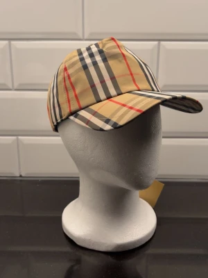 Rutig Burberry keps i beige och svart - Snygg keps från Burberry med klassiskt rutigt mönster i beige, svart, vitt och röda detaljer. Justerbar baktill med svart spänne och Burberry London England-märke. Perfekt accessoar för dig som gillar ikonisk design och streetstyle.