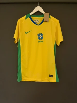 Brasil fotbollströja gul M - Säljer en Brasil fotbollströja i storlek M. Tröjan är gul med gröna och blå detaljer, har CBF-emblem på bröstet och Nike-logga. Tillverkad i lätt polyester med Dri-Fit-teknologi för skön känsla under matchen. Perfekt för dig som älskar fotboll och Brasilien. Det står vini Jr där bak med nummer 7