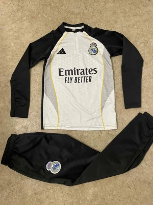 Real Madrid träningsset Adidas - Snyggt Real Madrid träningsset från Adidas med långärmad tröja och matchande byxor. Tröjan är vit med svarta ärmar, gula detaljer och klubbmärke på bröstet, samt halv dragkedja. Byxorna är svarta med klubbmärke på benet. Perfekt för fotbollsträning.