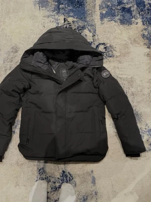 Svart MacMillan Parka från Canada Goose - Säljer en svart MacMillan Parka från Canada Goose Black Label. Jackan har huva, broderad logga på ärmen och dragkedja framtill. Den är quiltad och har en clean, stilren look med normal passform. Perfekt för kalla vinterdagar.