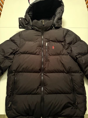 Svart pufferjacka från Polo Ralph Lauren - Svart pufferjacka från Polo Ralph Lauren med huva, två stora fickor med dragkedja och en röd broderad logga på bröstet. Jackan har grova dragkedjor och är perfekt för kalla dagar. Materialet är polyester och dun, vilket gör den både varm och lätt.