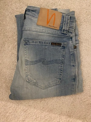 Ljusblå  jeans från Nudie Jeans -  Helt oanvända köpte online för 900 och de va för små för mig.  Säljer ett par ljusblå  jeans från Nudie Jeans med klassisk femficksdesign och snygga slitningar. Jeansen har en normal passform och är tillverkade i mjukt denimtyg. Perfekta för dig som gillar en avslappnad men stilren look.
