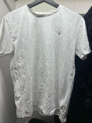 Vit t-shirt från GANT - Vit t-shirt från GANT med liten broderad logga på bröstet. Klassisk rund hals och korta ärmar. Tillverkad i mjuk bomull som känns skön mot huden. Enkel och stilren design som funkar till många olika looks.