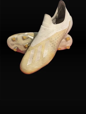 Adidas X 18+ elite  - Säljer ett par vita fotbollsskor från Adidas med guldiga och silverfärgade detaljer. Skorna har en strömlinjeformad design utan snörning och en platt sula med fasta dobbar för bra grepp på planen. Ovandelen är tillverkad i syntetmaterial med meshstruktur för lätthet och ventilation. Skorna är i strl 44 2/3 och är i extremt bra skick!