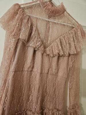 Ljusrosa långklänning i spets - Romantisk ljusrosa långklänning i spets med volanger och hög krage. Klänningen har långa ärmar och flera lager volangdetaljer längs kjolen och ärmsluten. Transparent över axlar och ärmar för en extra elegant look.