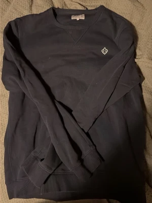 Mörkblå sweatshirt från Morris - Mörkblå sweatshirt från Morris med broderad vit logga på bröstet. Tröjan har rund hals, långa ärmar och ribbade muddar vid ärmslut och nederkant. Tillverkad i mjuk bomull som är skön att ha på sig. Perfekt för chill dagar eller när du vill ha en enkel stil.