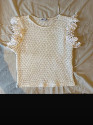 Vit topp med spetsvolang från Zara - Säljer en vit topp från Zara med korta ärmar och snygga spetsvolanger på axlarna. Toppen har ett stickat mönster och är croppad i modellen. Perfekt för dig som gillar romantiska detaljer och vill ha något som sticker ut.