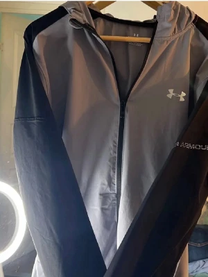 Grå och svart vindjacka Under Armour - Snygg vindjacka från Under Armour i grått och svart med huva och hel dragkedja framtill. Jackan har logga på bröstet och diskret Under Armour-tryck på ärmen. Perfekt för träning eller chill dagar ute. Lätt och smidig polyester som andas. Byten tas emot 