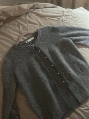 Grå kofta från Zara med pärlknappar - Mysig grå kofta från Zara i storlek S. Koftan har lång ärm, rund hals och är stickad i ett mjukt material. Framtill sitter snygga pärlknappar som ger en extra touch. Perfekt att slänga på sig när det är lite kyligt ute.