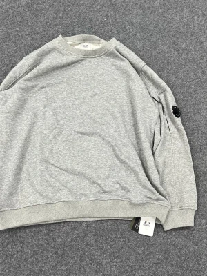 Grå sweatshirt från C.P. Company - Säljer en stilren grå sweatshirt från C.P. Company med klassisk rund hals och ribbade muddar. Tröjan har en cool detalj med en svart lins på ärmen och är tillverkad i mjuk bomull. Perfekt för dig som gillar enkel och snygg streetwear.