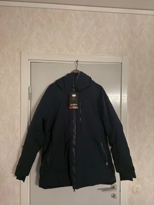 Svart vindjacka från CMP med huva - Snygg svart vindjacka från CMP med huva och hel dragkedja framtill. Jackan har två sidofickor med dragkedja och en stilren design som passar perfekt för blåsiga dagar. Materialet är slitstarkt och jackan har en normal passform.