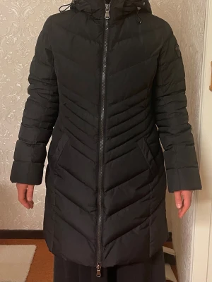 Svart lång pufferjacka från Pajar Canada  - Säljer en svart, lång pufferjacka från Pajar Canada med huva och snygga quiltade detaljer. Jackan har tvåvägsdragkedja framtill, sidofickor och extra dragkedjor vid sidorna för mer rörelsefrihet. Perfekt för kalla dagar och har en stilren look som funkar till allt.