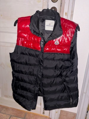 Moncler dunväst röd och svart XXL - Säljer en fet dunväst från Moncler i storlek XXL. Västen har en schysst färgblockad design med röd upptill och svart nedtill, samt Moncler-logga på bröstet. Materialet är glansigt och quiltat med dragkedja framtill och fickor med dragkedja.