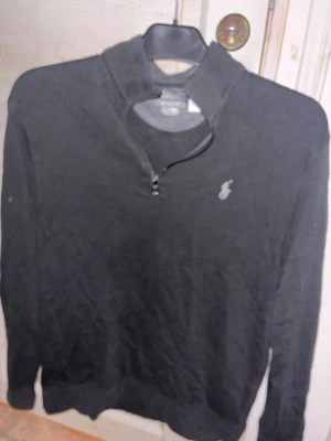 Svart half zip tröja från Polo Ralph Lauren - Svart långärmad tröja från Polo Ralph Lauren med half zip och klassisk logga på bröstet. Tröjan har en stilren design med ståkrage och är tillverkad i mjuk bomull. Perfekt för dig som gillar enkel och snygg stil.