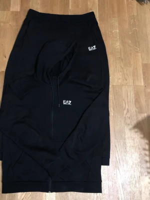 EA7 Tracksuit - EA7 Emporio Armani tracksuit – svart Stilren och bekväm tracksuit från EA7 Emporio Armani. Set med zip-hoodie och matchande byxor, båda med vit EA7-logga. Mycket fint skick, sparsamt använd och utan fläckar, hål eller slitage. Mjukt och följsamt material med sportig passform. Passar perfekt till vardag, resor och streetwear. Rök- och djurfritt hem. Skickas snabbt.