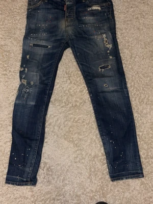 Dsquared2 Jeans ”Cool Guy” - Säljer ett par blå skinny jeans från Dsquared2 med coola slitningar, hål och färgstänk på både fram- och baksida. Jeansen har klassisk femficksdesign och är tillverkade i bomull. Perfekt för dig som gillar en edgy streetstil.