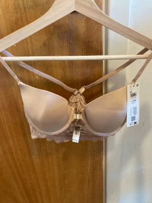 Bh med spetsdetaljer 75B - Snygg T-shirt bh i beige från Lindex med vadderade kupor och spetsdetaljer bak. Axelbanden är korsade och justerbara för extra stöd. Spetsen har ett blommigt mönster och ger en romantisk vibe. Perfekt för dig som gillar mjuka färger och fina detaljer.Endasr provad.Slutsåld på hemsidan.