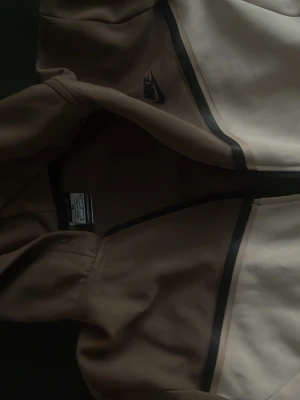 Nike beige och vit vindjacka - Nike vindjacka i beige och vitt med svarta detaljer och dragkedja framtill. Jackan har en sportig look med långärmade ärmar och stilrena linjer. Perfekt för dig som gillar streetwear och vill ha en clean jacka med snygg design.