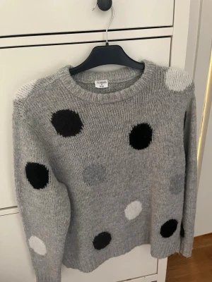 Grå stickad prickig tröja Stories - Mysig grå stickad tröja från Stories i storlek S, dekorerad med stora svarta, vita och grå prickar. Rund halsringning och långa ärmar. Perfekt för dig som gillar lekfulla detaljer och vill ha en skön tröja till hösten, våren eller vintern.