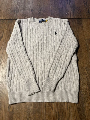 Ljusgrå kabelstickad tröja Polo Ralph Lauren - Säljer en ljusgrå kabelstickad tröja från Polo Ralph Lauren med klassisk rund hals och broderad logga på bröstet. Tröjan har ribbade muddar vid ärmslut och nederkant samt långärmad passform. Perfekt för kyliga dagar och stilren look.