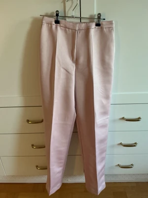 Ljusrosa kostymbyxor med rak passform - Jag säljer ett par  ljusrosa kostymbyxor med rak siluett och hög midja. Byxorna har pressveck framtill och sidfickor. Materialet är polyester och är lite glansigt. Köparen står för frakten