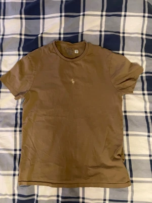Brun t-shirt från Polo Ralph Lauren - Snygg brun t-shirt från Polo Ralph Lauren med klassisk rund hals och korta ärmar. Ikoniska Polo-loggan broderad på bröstet i beige. Tillverkad i mjuk bomull som känns skön mot huden. Perfekt för en chill och stilren look.