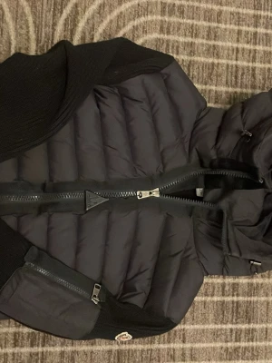 Svart pufferjacka från Moncler - Säljer en svart pufferjacka från Moncler med stickade partier på ärmar och rygg. Jackan har en stor dragkedja framtill, Moncler-logga på ärmen och quiltad design på framsidan. Perfekt för dig som vill ha en snygg och varm jacka med coola detaljer.